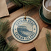 A circular ornament with the text " Cayman Brac Cayman Islands" printed on it, surrounded by pine branches and a cup of coffee on a wooden surface.