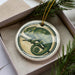 A ceramic ornament in the shape of a chameleon, with a green and brown color scheme, is displayed in a box with pine branches.