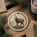 a decorative ornament with a deer illustration on a wooden surface, accompanied by a cup of coffee and some pine branches.