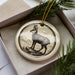A ceramic ornament in the shape of a deer, with a detailed illustration of the animal on a circular base, is displayed in a box with pine branches.