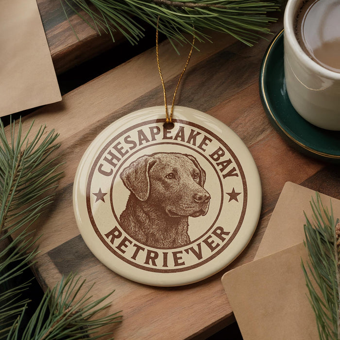 A round ornament featuring a brown dog's head, with the text "CHESAPEAKE BAY RETRIEVER" printed on it, is displayed on a wooden surface alongside a cup of coffee and some pine branches.