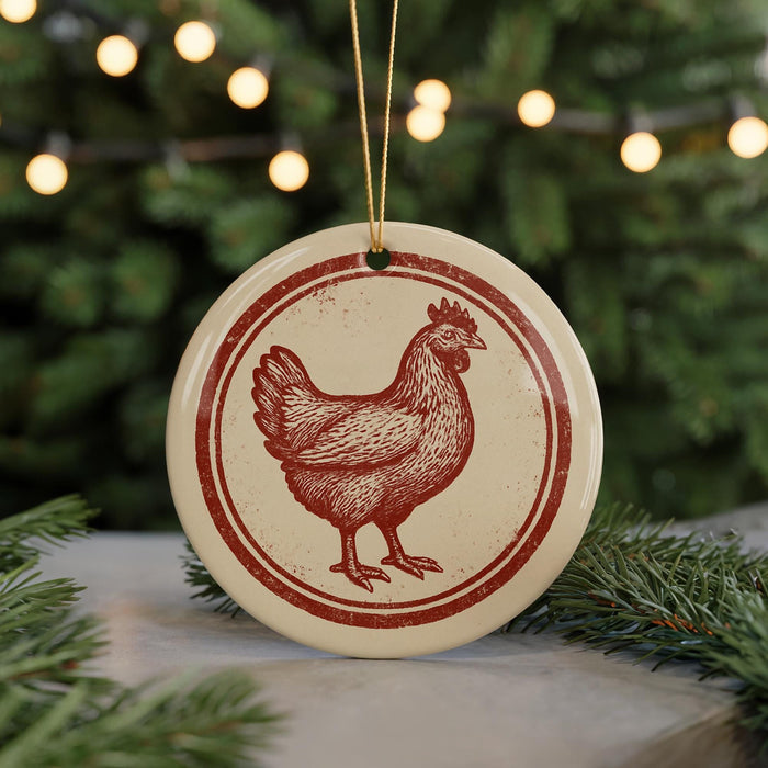 A round ornament featuring a red rooster design is hanging from a string, surrounded by pine branches and twinkling lights.