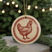 A round ornament featuring a red rooster design is hanging from a string, surrounded by pine branches and twinkling lights.