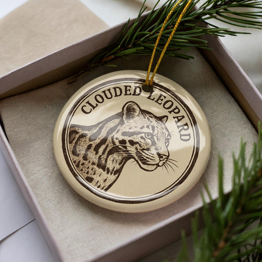 A circular ornament featuring a leopard's head, with the text "CLOUDED LEOPARD" printed on it, is displayed in a box surrounded by pine branches.