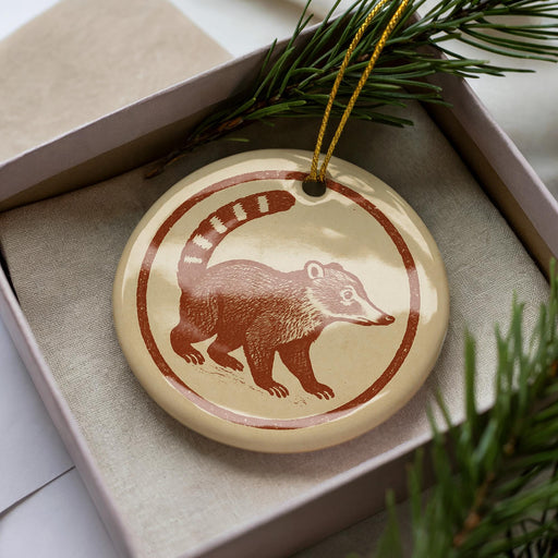 A round ornament featuring a brown raccoon or squirrel on a beige background, placed in a box with pine branches.
