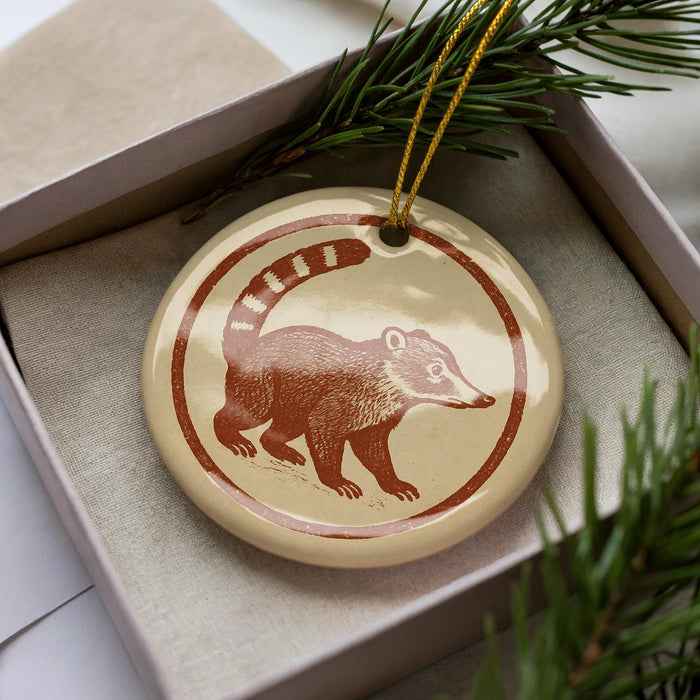 A round ornament featuring a brown raccoon or squirrel on a beige background, placed in a box with pine branches.