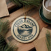 A circular ornament with the text "COEUR D'ALENE IDAHO * USA" printed on it, surrounded by pine branches and a cup of coffee on a wooden surface.