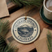 A decorative ornament featuring the text "COLORADO SPRINGS COLORADO USA" surrounded by a mountain illustration, placed on a wooden surface with pine branches and a cup of coffee nearby.