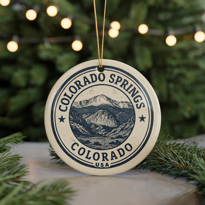 A round ornament with the text "COLORADO SPRINGS COLORADO USA" and a mountain illustration, hanging from a string against a blurred background of Christmas lights.