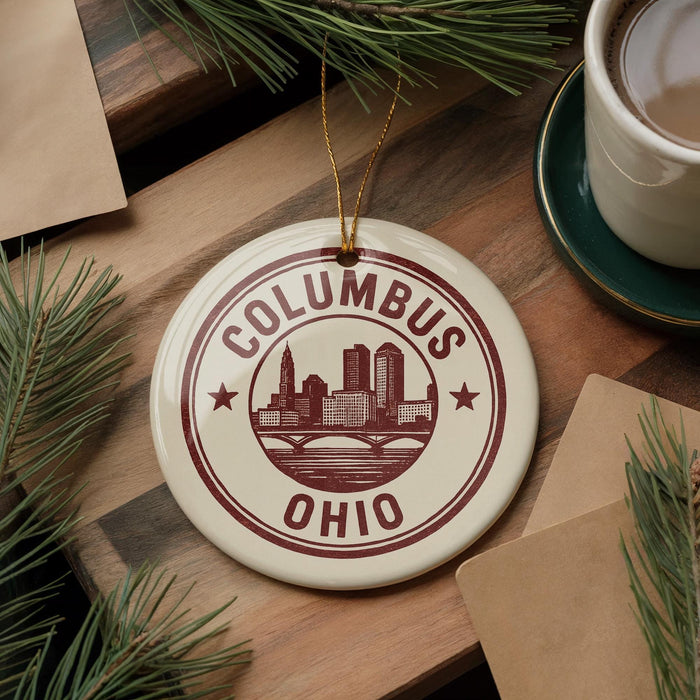 A ceramic ornament with the text "COLUMBUS OHIO" and a graphic of a city skyline, placed on a wooden surface alongside pine branches and a cup of coffee.