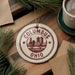 A ceramic ornament with the text "COLUMBUS OHIO" and a graphic of a city skyline, placed on a wooden surface alongside pine branches and a cup of coffee.