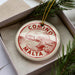 A circular ornament with the text "COMINO MALTA" printed on it, placed inside a box with pine branches.