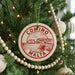 A Christmas tree decorated with ornaments, including a circular ornament with the text "COMINO MALTA" and a wooden bead decoration.