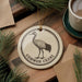 a decorative ornament with a black and white illustration of a crane, placed on a wooden surface. The ornament has the text "COMMON CRANE" printed on it.
