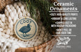 Ceramic ornaments, featuring a bird and text, are displayed in a basket with greenery.