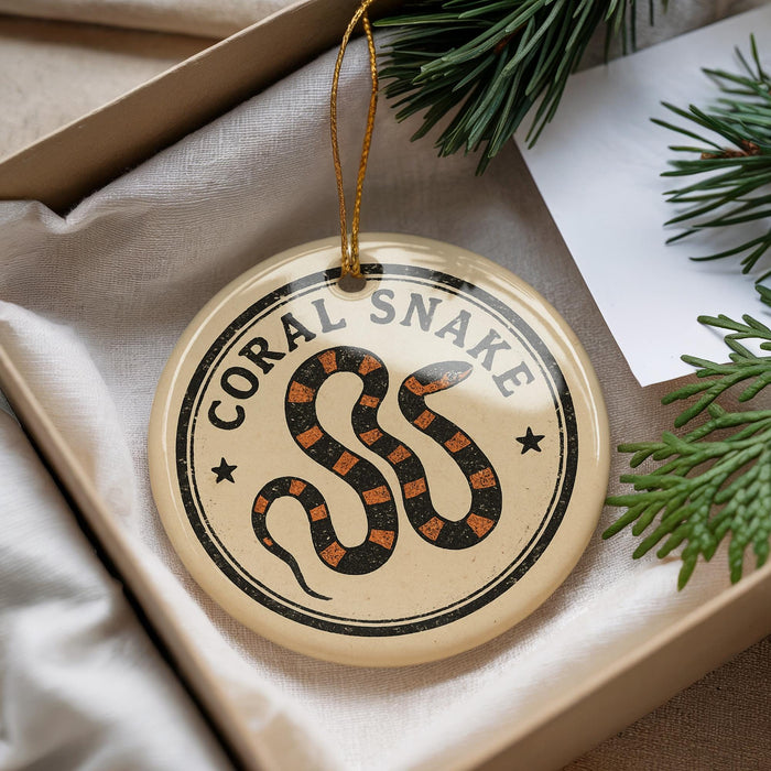 A round ornament with a black and orange snake design, hanging from a string, placed inside a box with pine branches.
