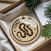 A round ornament with a black and orange snake design, hanging from a string, placed inside a box with pine branches.
