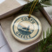 A circular ornament with the text "CORFU GREECE" and a graphic of a building and mountains, hanging from a string.