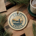 A circular ornament with the text "CORFU GREECE" and an illustration of a coastal town, surrounded by pine branches and a cup of coffee on a wooden surface.
