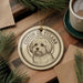 A circular ornament featuring a dog's portrait is displayed on a wooden surface, surrounded by pine branches and a cup of coffee.
