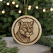A round ornament featuring a close-up of a cougar's face, with a blurred background of a Christmas tree and twinkling lights.