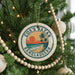A circular ornament on a Christmas tree, featuring a tropical beach scene with palm trees and a sunset.