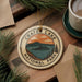 A circular ornament with the text "Crater Lake National Park" and an illustration of a lake surrounded by mountains, pine trees, and a sunset sky.