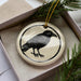 A ceramic ornament in the shape of a black bird is displayed in a box, surrounded by pine branches.