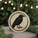 A ceramic ornament featuring a black crow silhouette is displayed against a blurred background of Christmas lights and pine branches.