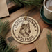 A round ornament featuring a wolf's head, with the text "CZECHOSLOVAKIAN WOLFdog" printed on it, is displayed on a wooden surface alongside a cup of coffee and some pine branches.