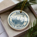 A decorative ornament in a box, featuring a circular design with a cityscape and text, surrounded by pine branches.