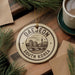 A decorative ornament with the text "DAEJEON SOUTH KOREA" on it, surrounded by pine branches and a cup of coffee on a wooden surface.