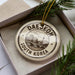 A decorative ornament in a box, featuring a circular design with text and an illustration of a cityscape.