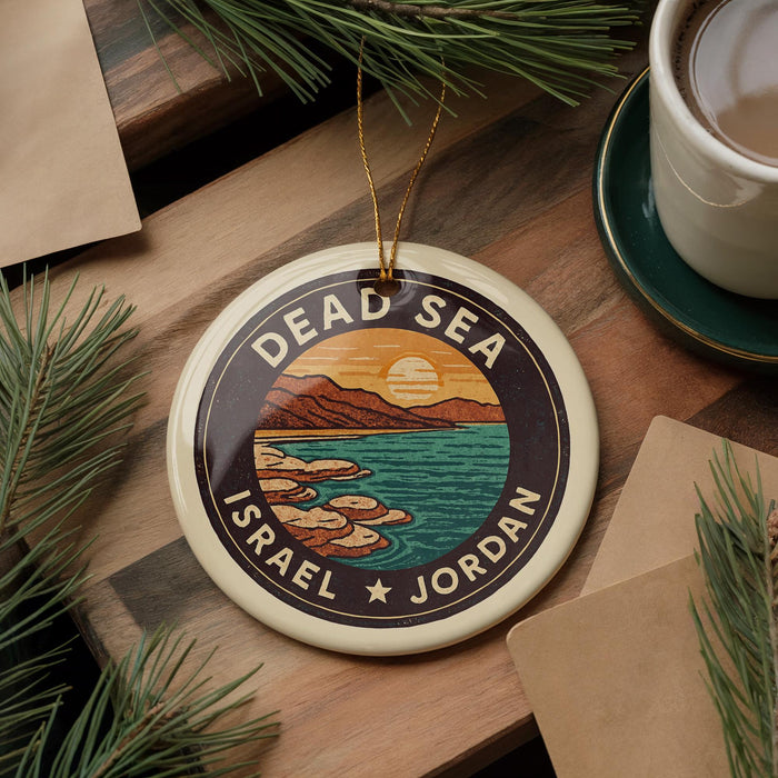 A decorative ornament featuring a circular design with a sunset over a body of water, surrounded by mountains and islands, is displayed on a wooden surface alongside a cup of coffee and some pine branches.