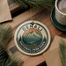 A decorative ornament featuring the name "DENALI" and the text "NATIONAL PARK PARK U.S.A." is displayed on a wooden surface, surrounded by pine branches and a cup of coffee.