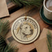 A circular ornament featuring a dog's head and the text "DOGO ARGENTINO" is displayed on a wooden surface, surrounded by pine branches and a cup of coffee.