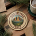 A decorative ornament with the text "DOLOMITES ITALY" and a mountain landscape design, placed on a wooden surface next to a cup of coffee and pine branches.