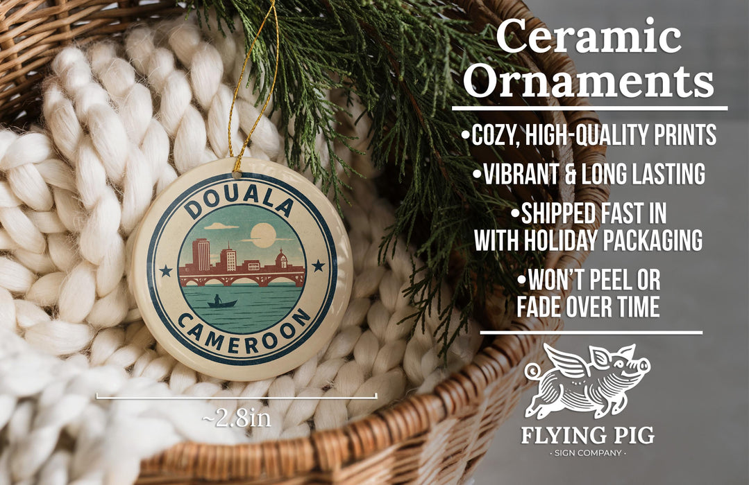 Ceramic ornaments, specifically a DOUALA CAMERON ornament, are displayed in a woven basket. The ornament is described as cozy, high-quality prints, vibrant, and long lasting, and it is shipped fast in holiday packaging. The ornament is not peeling or fading over time. The image also includes a scale indicating the ornament's size, with the text "2.8in" and a logo for "FLYING PIG - SIGN COMPANY".