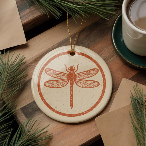 a ceramic ornament with an orange dragonfly design, placed on a wooden surface alongside a cup of coffee and some pine branches.