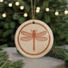 A ceramic ornament featuring a detailed illustration of a dragonfly is displayed on a table, with a blurred background of a Christmas tree and twinkling lights.