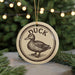 A ceramic ornament featuring a duck, with the word "DUCK" written on it, is displayed in front of a blurred Christmas tree with twinkling lights.