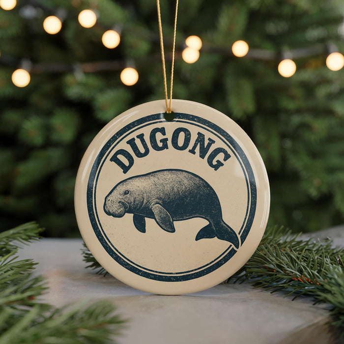 A round ornament with the word "DUGONG" printed on it, featuring a graphic of a dugout or dugout-like structure. The ornament is hanging from a string, and there are pine branches visible in the background.
