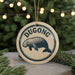 A round ornament with the word "DUGONG" printed on it, featuring a graphic of a dugout or dugout-like structure. The ornament is hanging from a string, and there are pine branches visible in the background.