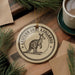 A circular ornament featuring a kangaroo, with the text "EASTERN GRAY KANGAROO" around the edge, is displayed on a wooden surface alongside a cup of coffee and some pine branches.