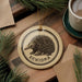 a circular ornament with a depiction of an echidna, a type of insect, and the word "ECHIDNA" written on it. The ornament is placed on a wooden surface, surrounded by pine branches and a cup of coffee.