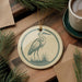 a round ornament with a bird illustration, placed on a wooden surface next to a cup of coffee and some pine branches.