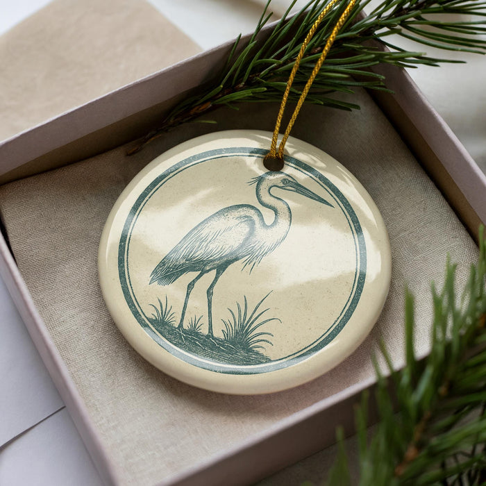 A ceramic ornament in the shape of a heron, with a green and white design, is displayed in a box with pine branches.