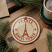 A circular ornament with the Eiffel Tower design, surrounded by pine branches and a cup of coffee on a wooden surface.