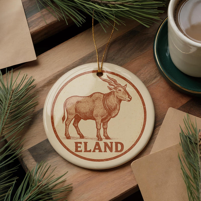A round ornament featuring a brown antelope, with the word "ELAND" written on it, is displayed on a wooden surface alongside a cup of coffee and some pine branches.