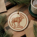 A round ornament featuring a brown antelope, with the word "ELAND" written on it, is displayed on a wooden surface alongside a cup of coffee and some pine branches.